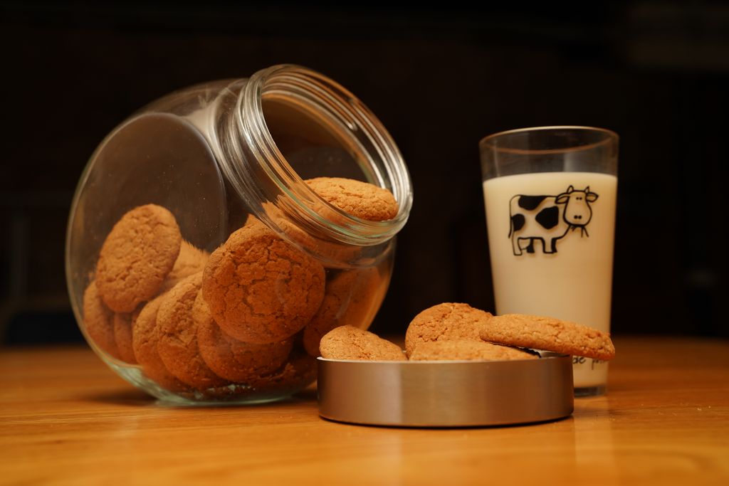 A glass jar spilling cookies next to a glass of milk on a wooden table, perfect for cozy kitchen vibes.